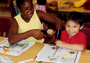 Two children of differing ages and ethnicities help each other to create milagos, small metal objects representing their hopes and dreams.