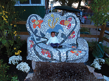 Surrounded by flowers, a toddler sits on an oversized tile mosaic "couch" in COSACOSA's North Philadelphia Healing Garden.